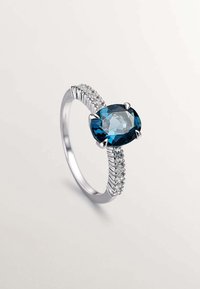 Anillo de plata con una piedra azul ovalada y facetada, engastada en cuatro garras, acentuado por pequeñas piedras claras a lo largo de la banda.