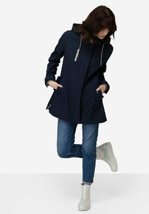 Marineblaue, wasserdichte Jacke mit Kapuze, ausgestattet mit zwei Taschen vorne und einem hellgrauen Reißverschluss. Kombiniert mit blauen Jeans und schimmernden Gummistiefeln.