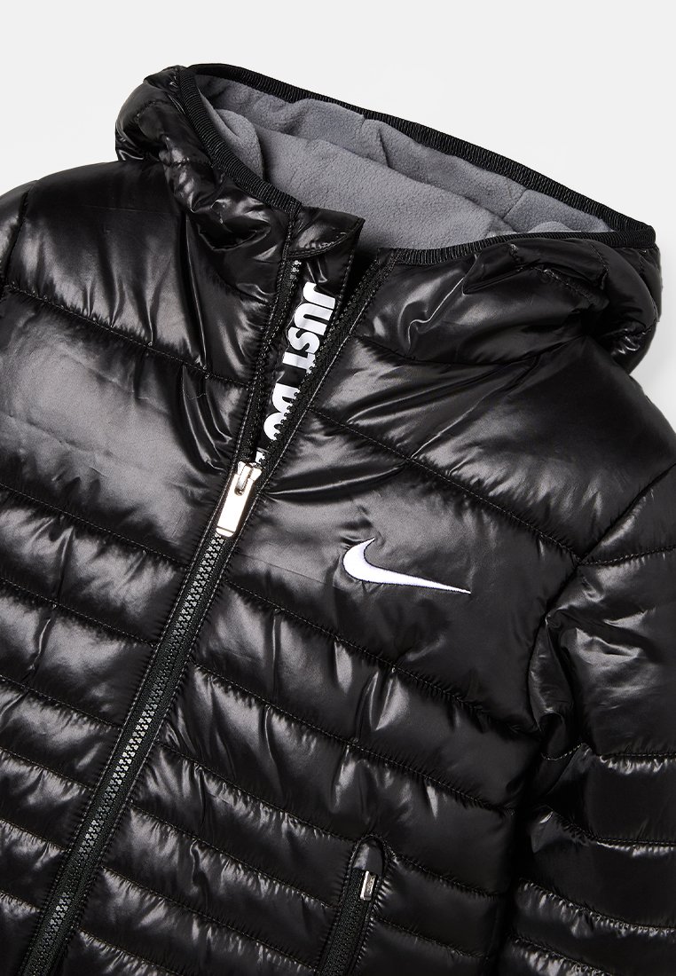 Doudoune noire avec une texture brillante, design matelassé, capuche et zip argenté. Présente un logo Nike blanc et une étiquette "JUST DO IT" à l'intérieur.