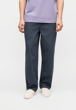 Osoba stojaca pred jednofarebným pozadím, oblečená v voľných tmavosivých nohaviciach, svetlofialovej košeli a krémovobielych slip-on topánkach.