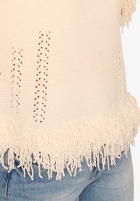 Cremfarbener Strickoberteil mit offenen Webakzenten an der Vorderseite und einem fransigen Saum. Der Stoff wirkt weich und texturiert.