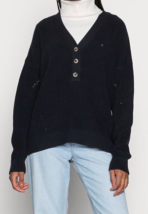 Person trägt einen schwarzen Strickpullover mit V-Ausschnitt und drei Knöpfen, darunter einen weißen Rollkragenpullover und hellblaue Jeans.