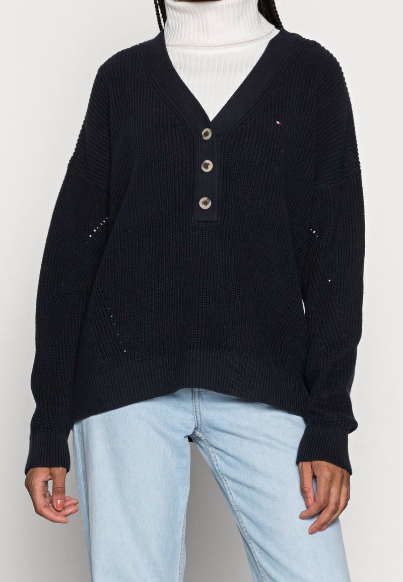 Personne portant un pull en maille noir à col en V avec trois boutons, un col roulé blanc en dessous et un jean bleu clair.