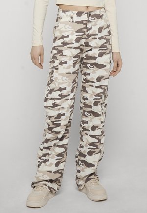 Persona che indossa pantaloni cargo mimetici beige, marroni e bianchi con multiple tasche e sneakers beige chiare su uno sfondo semplice.