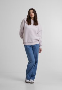 Lys rosa sweatshirt med rund hals og brodert logo, kombinert med høy midje blå jeans og hvite sneakers. Enkelt bakgrunn.