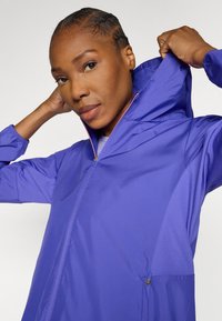 Μωβ αδιάβροχο με κουκούλα, διαθέτει φερμουάρ μπροστά, υφασμάτινα πάνελ με υφή και ελαστικά μανίκια. Λείο υλικό με εφαρμοστή σχεδίαση.