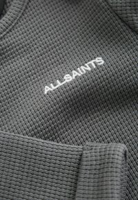 Pullover aus gewaschenem Waffle-Strick in Anthrazitgrau mit Rundhalsausschnitt und einem auf der Brust gestickten weißen "ALLSAINTS"-Logo. Weiche Textur mit gerippten Bündchen.