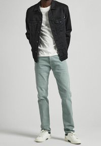 Giacca di jeans nera con due tasche sul petto, t-shirt bianca, jeans verde chiaro e sneakers bianche. L'outfit presenta una vestibilità casual e linee pulite.