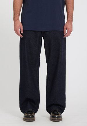 Homme portant un jean bleu foncé ample, des chaussures noires à semelles épaisses et une chemise bleu marine, debout sur un fond uni.