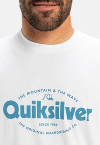Bílý bavlněný tričko s velkým modrým logem Quiksilver, doplněné textem a grafikou vlny, kulatým výstřihem, krátkým rukávem a volným střihem.