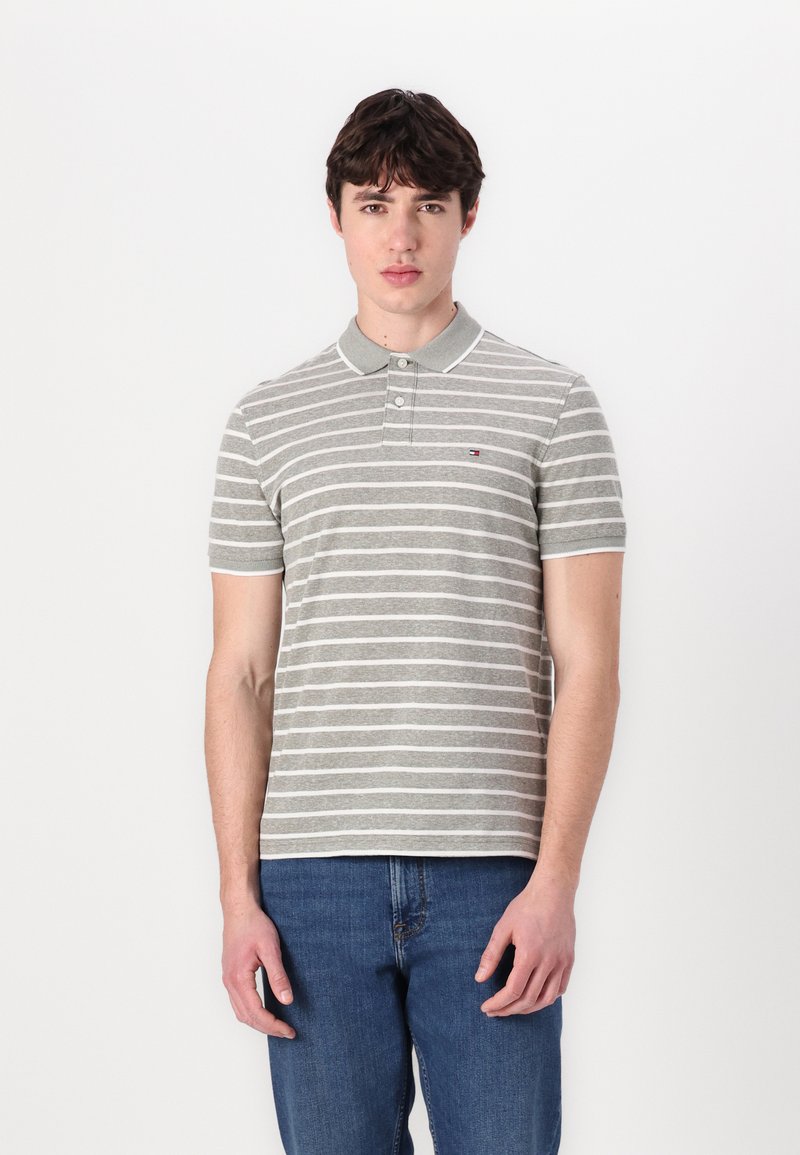 Γκρι και λευκή ριγέ πικέ μπλούζα με γιακά, που διαθέτει ένα μικρό λογότυπο στο στήθος, συνδυασμένη με μπλε τζιν παντελόνι.