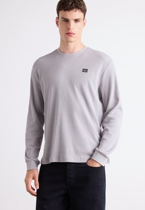Gray long-sleeve shirt made of textured fabric, featuring a round neckline and a small logo patch on the left chest. Dark pants complete the look.