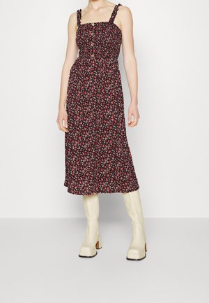 Robe midi noire à imprimé floral avec corsage froncé, encolure carrée et bretelles tressées, associée à des bottes crème montantes jusqu'au genou et des talons blocs.