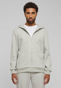 Lys grå zip-up hoodie lavet af blødt stof, med hætte, to forlommer og ribbede manchetter. Bar over en hvid skjorte.