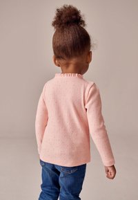 Hele roosa pika varrukaga särk, millel on kõrgendatud täpid ja volangidega kaelus. Sobitatud siniste teksapükstega. Vaade tagant.