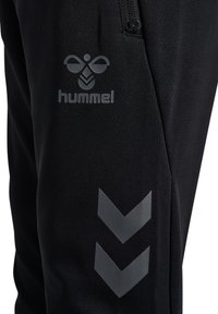 Svarta träningsbyxor med en slät tygkvalitet, Hummel-logotyp i grått och reflektiv chevron-detalj på sidan.
