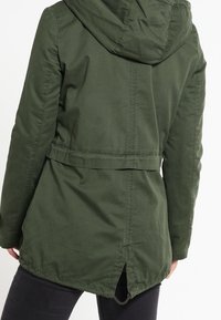 Chaqueta de longitud media de color verde oscuro, confeccionada con un material ligero, que cuenta con capucha, cintura ajustable con cordón y dobladillo trasero ventilado.