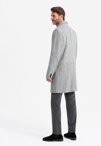 Abrigo de lana gris con un corte entallado, cuello con muesca y tres botones en los puños, llevado sobre pantalones gris oscuro y botines negros.