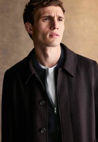 Czarny wełniany płaszcz z kołnierzykiem i zapięciem na guziki. Nosi się go na granatowej kurtce na zamek i białej koszulce, o gładkiej teksturze i dopasowanym kroju.