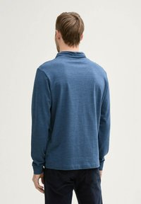 Niebieska, długozasłonięta koszulka polo z fakturowanej tkaniny, z luźnym kołnierzykiem i prostym dołem, noszona przez osobę widzianą z tyłu.