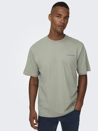 Lichtgroene katoenen T-shirt met korte mouwen, relaxed fit en geborduurde tekst op de linkerborst. Draagt met donkere denim jeans.