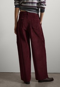Pantaloni a gamba larga in velluto a coste bordeaux con due tasche posteriori e tasche laterali cargo. Texture morbida con dettaglio a righe verticali, abbinati a scarpe scure.
