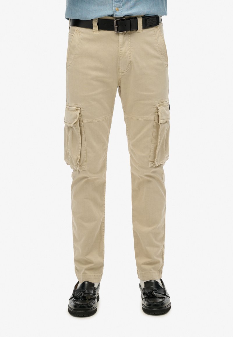 Beige cargo broek met meerdere zijzakken en een rechte pijp ontwerp, gemaakt van een stevig materiaal. Draag met een zwarte riem en glanzende schoenen.