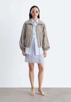 Kvinde iført beige lynlåstrøje, lyseblå skjorte med knapper, gennemsigtig lys lilla nederdel, hvide solbriller og hvide sandaler med stropper, står på gråt gulv.