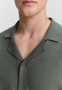 Närbild av en persons nacke och nedre ansikte som bär en texturerad, olivgrön skjorta med krage och en enda knapp knäppt.