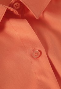 Camicia button-up corallo con una texture liscia, dotata di colletto a punta e bottoni rotondi abbinati. La ripresa ravvicinata mostra le cuciture dettagliate.