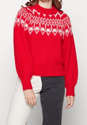 Pull en laine rouge avec un motif géométrique blanc sur les épaules, associé à un pantalon bicolore blanc et gris foncé.