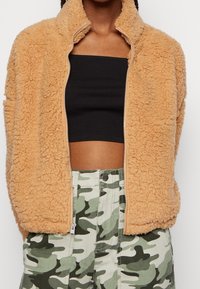 Veste en polaire beige avec col montant et fermeture éclair, portée sur un top court noir, associée à un pantalon camouflage vert. Texture douce visible.