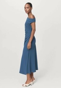 Blaues schulterfreies Kleid mit einem taillierten Oberteil und einem ausladenden Rock. Glattes Material mit dezenten Drapierungsdetails. Getragen mit weißen offenen Zehenschuhen.