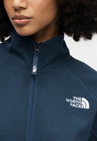 Σκούρο μπλε φλις μπουφάν με ψηλό γιακά, κλείσιμο με φερμουάρ και λευκό λογότυπο "The North Face" στο στήθος. Απαλή υφή και λείο ύφασμα.