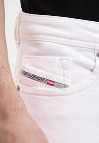 Gros plan d'une personne portant un jean en denim blanc avec une étiquette visible de la marque Diesel Industry sur la petite poche avant.