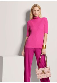 Leuchtend pinkes, geripptes Strickoberteil mit hohem Kragen und kurzen Ärmeln, kombiniert mit lebhaften pinken Hosen. Hält eine strukturierte pinke Handtasche mit goldenen Akzenten.