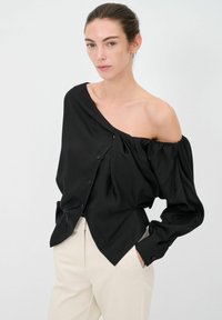 Blusa nera off-the-shoulder realizzata in tessuto liscio con chiusura a bottoni, polsini elasticizzati e un design drappeggiato, abbinata a pantaloni beige.