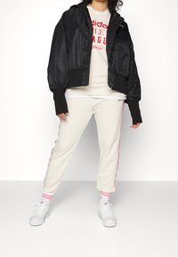 Zwart satijnen cropped jack met geribde manchetten, witte sweatshirt met rode tekst, crèmekleurige joggingbroek met rode zijstrepen en witte sneakers.