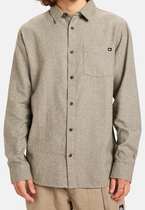 Beige langärmliges Hemd mit Knöpfen, linker Brusttasche und dunklen Knöpfen, kombiniert mit hellbrauner Hose.