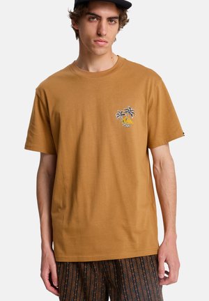 Jeune homme portant un t-shirt marron moutarde avec un petit dessin de palmier et de soleil sur la poitrine, ainsi qu'un pantalon à motifs, debout devant un fond blanc.