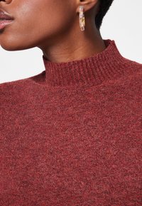 Primo piano di una persona che indossa un maglione a collo alto in maglia rossa e un orecchino ad anello traslucido multicolore all'orecchio sinistro.