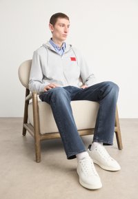 Grå hettejakke med rød logo, lagd over en blåstripet skjorte. Brukt sammen med blå jeans og hvite sneakers, sittende på en trebenk.