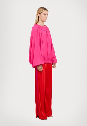 Blusa lunga a maniche corte di un rosa brillante con scollo arricciato, abbinata a pantaloni rossi a gamba larga con righe verticali sui lati. Texture liscia.