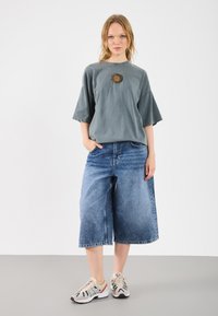 Szary, oversize'owy t-shirt z grafiką słońca; zestawiony z szerokimi, niebieskimi dżinsowymi kulotami; wykończony beżowymi i granatowymi trampkami.