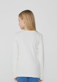 Biały sweter z długim rękawem o prążkowanej fakturze, posiada falbaniasty dół i jest zestawiony z niebieskimi dżinsami. Widok z tyłu.