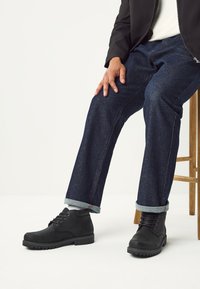 Schwarze Leder-Stiefeletten mit runden Zehen und Schnürsenkeln, kombiniert mit dunkelblauen Jeans mit umgeschlagenen Bündchen und sichtbaren Nahtdetails.