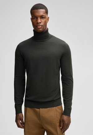 A man in a dark green turtleneck sweater stands against a light gray background. His expression is neutral, and he wears brown pants.