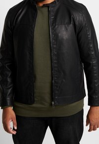 Zwarte leren jas met een rits aan de voorkant, opstaande kraag en twee voorzakken. Ggedragen over een groene textuurtrui en donkere jeans.
