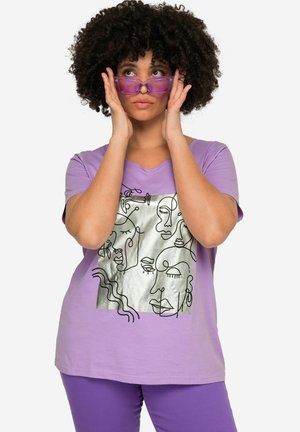Lila T-shirt van katoen met een metallic zilveren gezichtsgrafiek. Het ontwerp heeft abstracte lijnkunst. Model draagt paarse zonnebril.