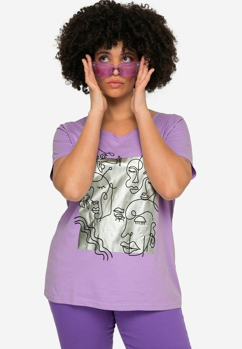 Lilla t-shirt lavet af bomuld med et metallisk sølv ansigt grafik. Designet har abstrakt linjekunst. Modellen bærer purpurfarvede solbriller.